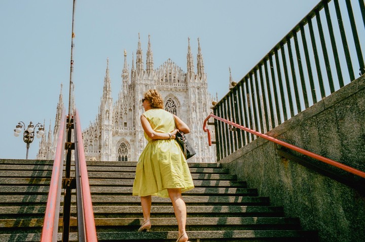 A woman in a yellow dress walks up a stone staircase with a large gothic cathedral in the background.