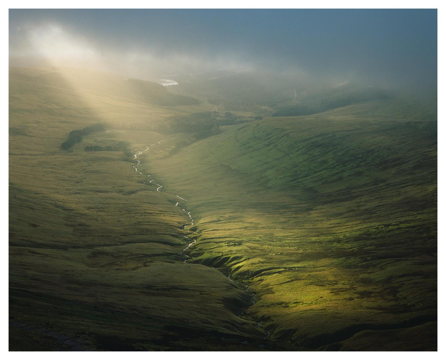 A winding river flows through a green valley under a hazy sky with a ray of light.