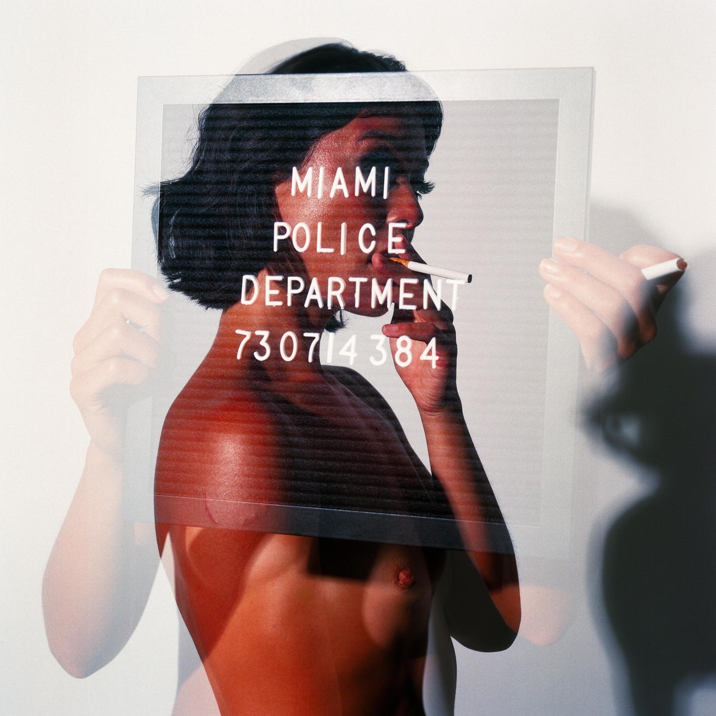 A shirtless woman with short dark hair holds a cigarette in her mouth while holding a clear sign that reads "MIAMI POLICE DEPARTMENT 730714384".