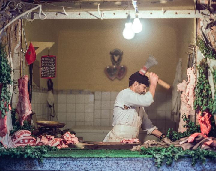 A man in a white apron and black hat chops meat with an axe in a butcher shop.