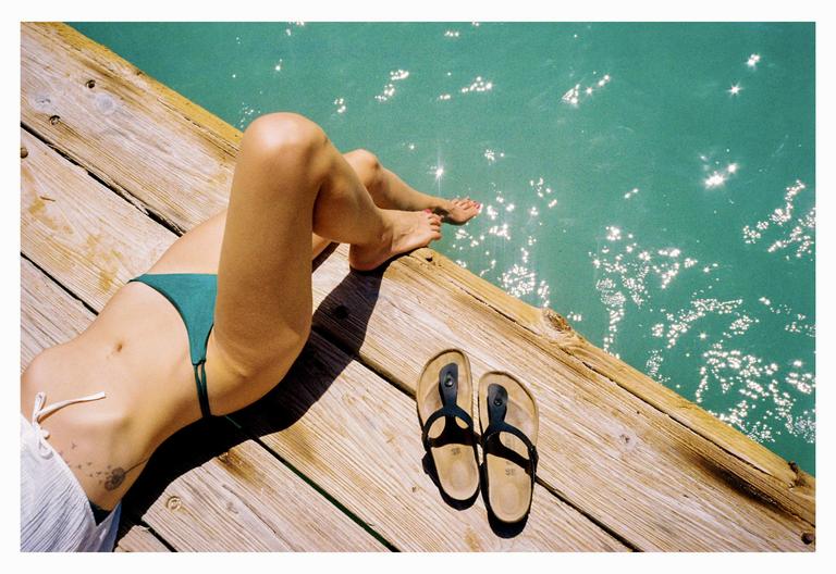 A woman in a bikini is lying on a wooden dock next to sparkling water, with a pair of sandals nearby.