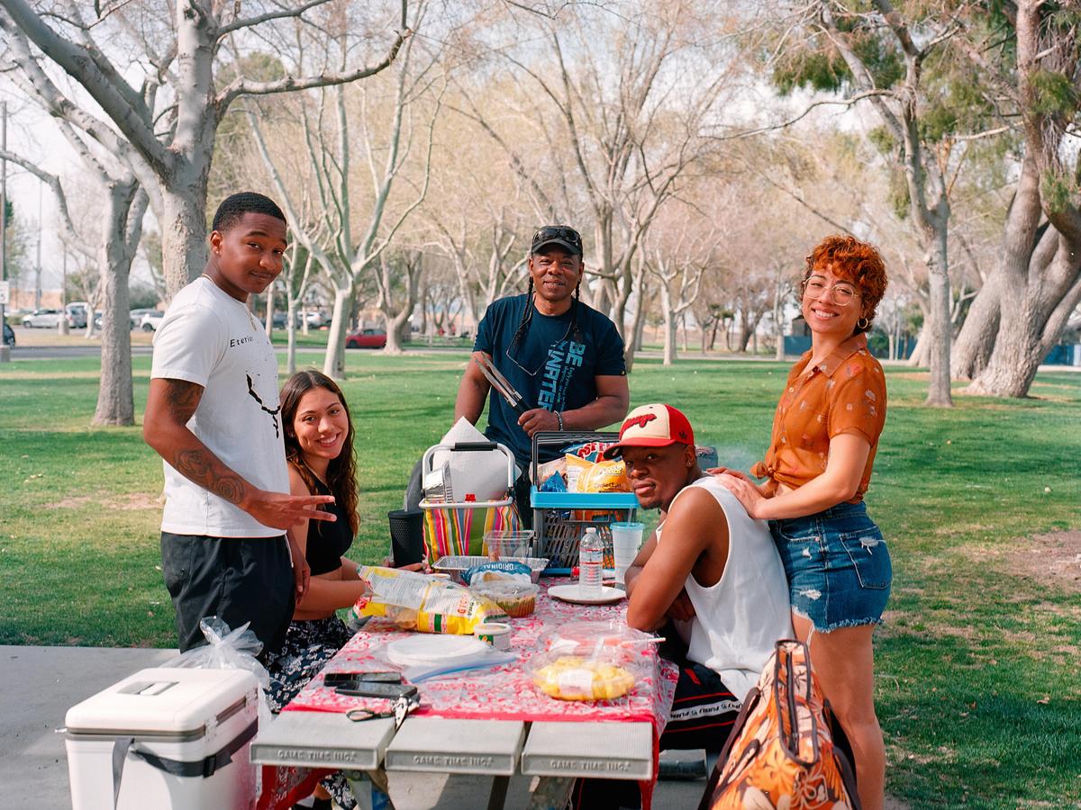 A group of five people are gathered around a picnic table with food and drinks in a park.