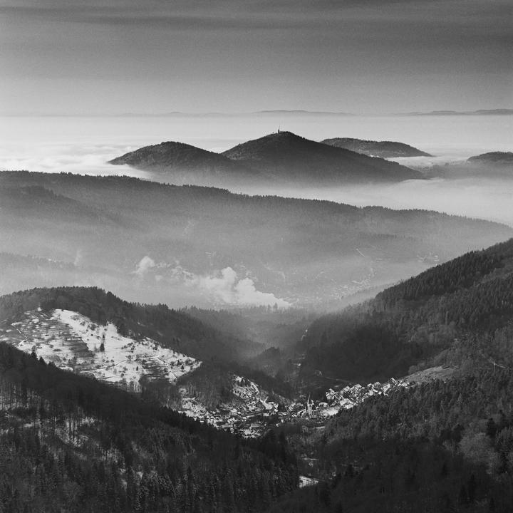 A village nestled in a valley is surrounded by forested mountains, with a sea of fog in the distance.