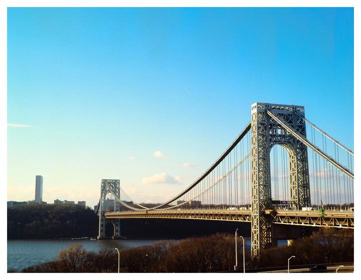 A suspension bridge spans a river with a city skyline and trees on the far bank under a clear sky.