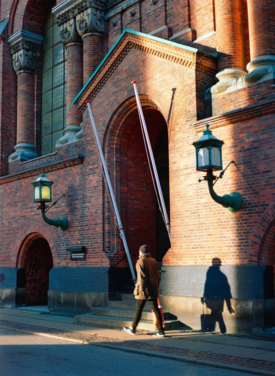 A man walks past a brick building with arched doorways and ornate lanterns, casting a long shadow on the sunlit wall.