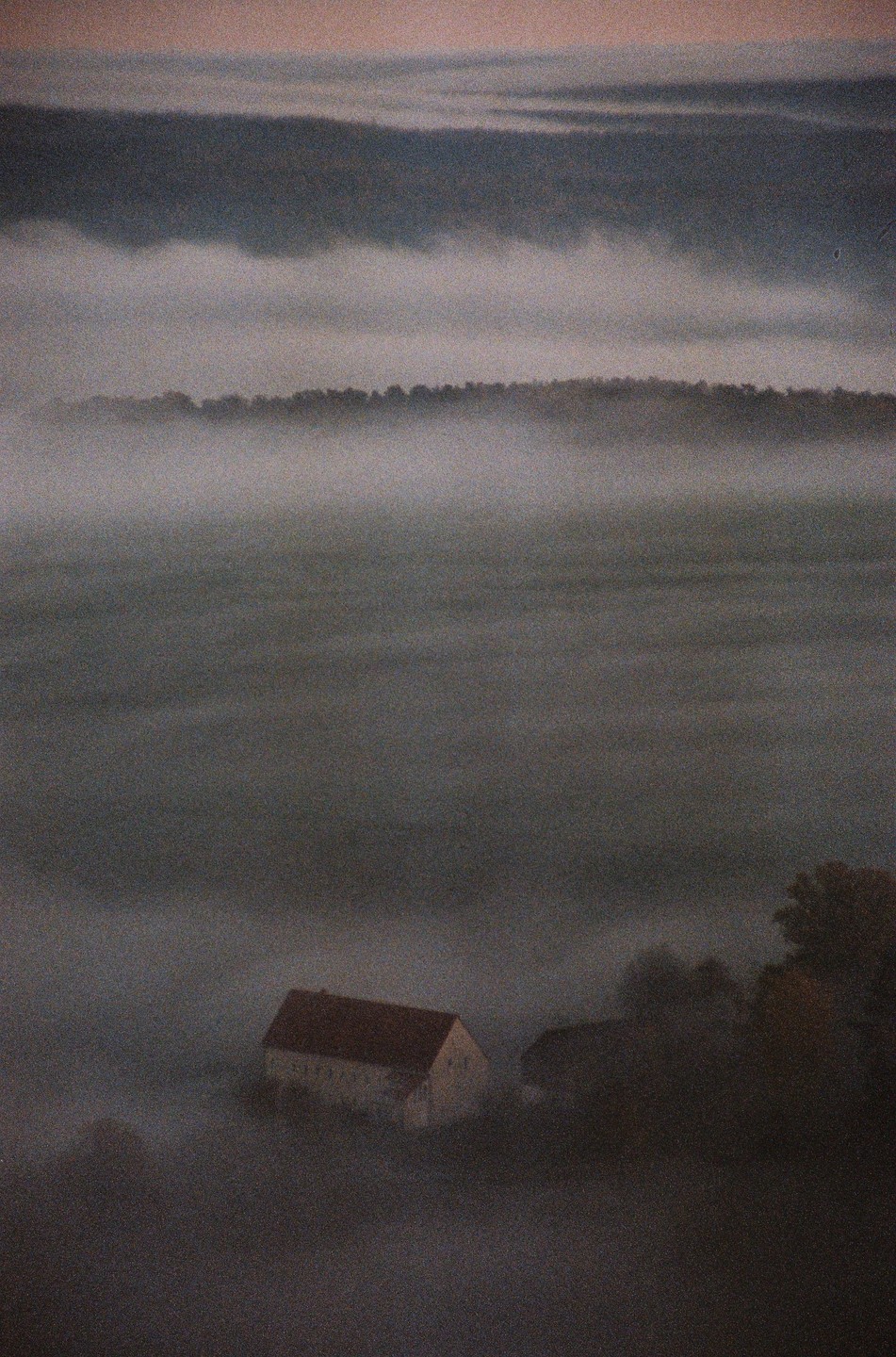 A house with a red roof is nestled in a valley filled with fog, with a line of trees visible in the distance.