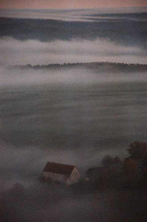 A house with a red roof is nestled in a valley filled with fog, with a line of trees visible in the distance.