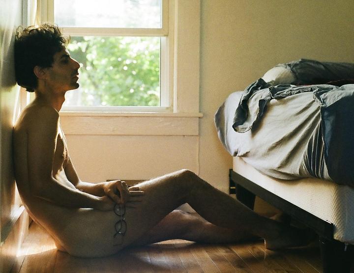 A man sits on a wooden floor next to a window and a bed, holding glasses in his hand.