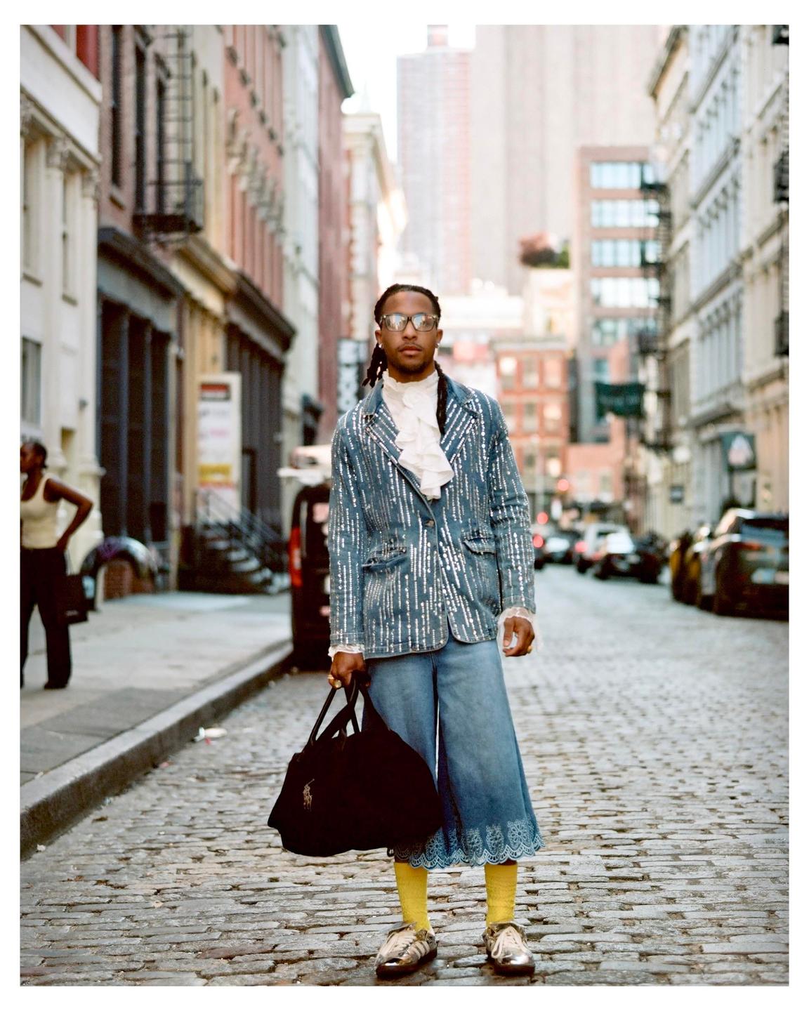 A man wearing a sequined jacket, culottes, and yellow socks stands on a cobblestone street holding a bag.