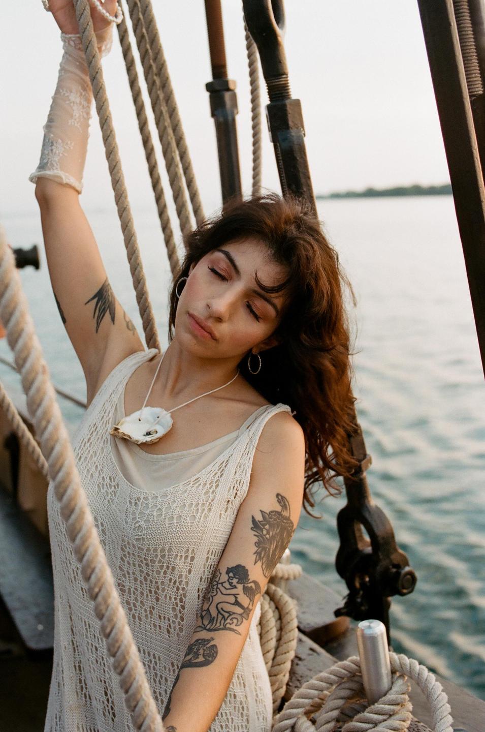 A woman with her eyes closed and arm raised is on a boat with ropes and water in the background.
