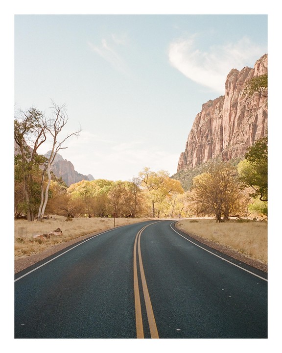 A winding road with double yellow lines leads through a valley with trees and a large rock formation under a bright sky.