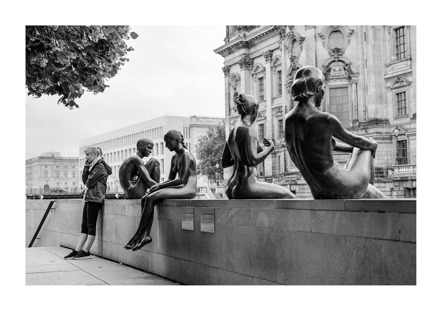 A girl stands next to a row of bronze statues of nude figures, with buildings in the background.