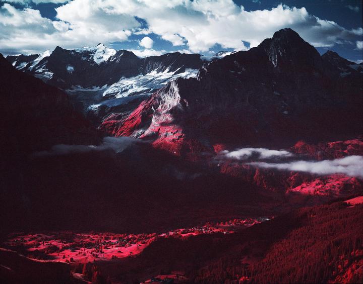 A mountain range with snow-capped peaks overlooks a valley with a small village and red foliage.