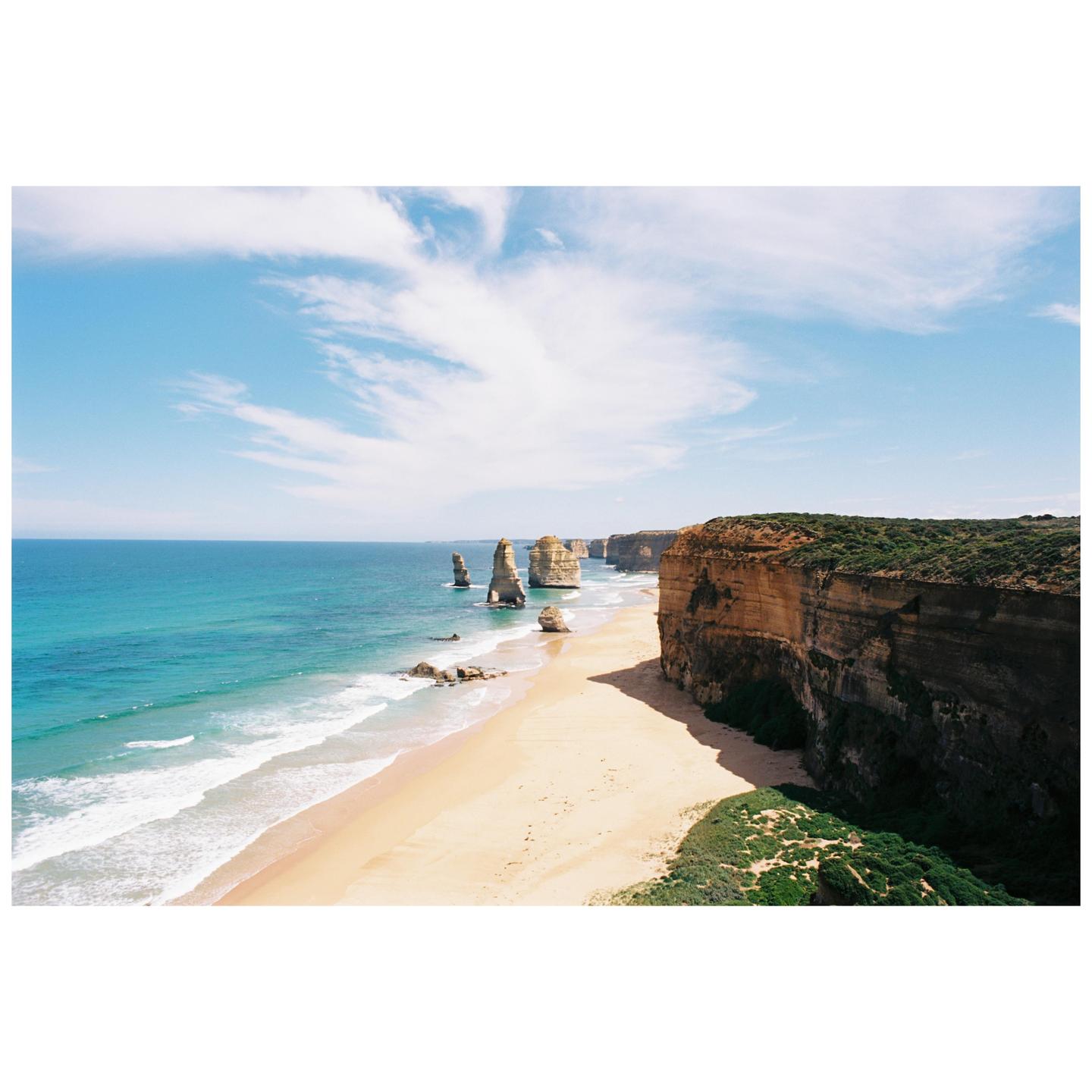A sandy beach with ocean waves is next to a tall cliff and several sea stacks under a blue sky with clouds.