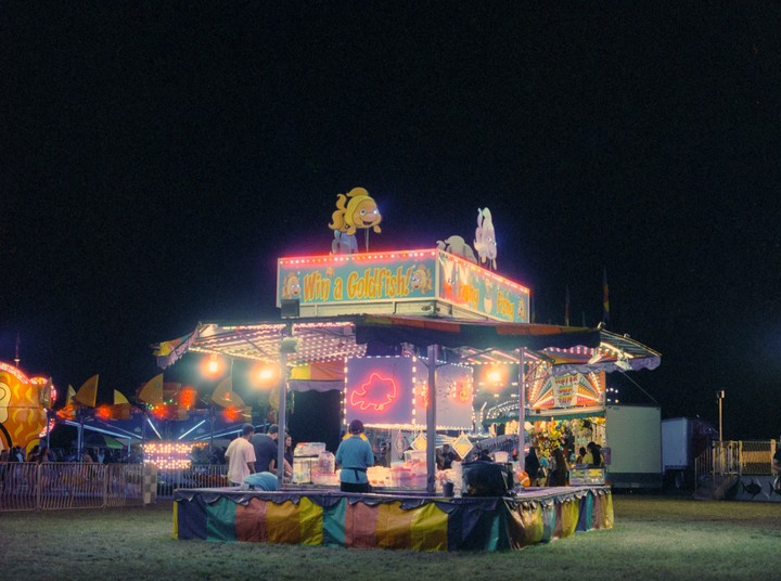 A carnival game booth with a "Win a Goldfish!" sign is brightly lit at night, with people gathered around it.