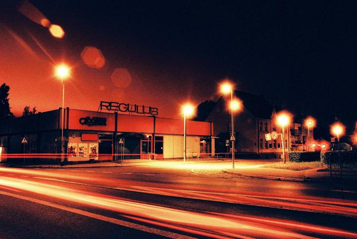 A street at night shows a building with the name "REGULUS" on it, with light trails from vehicles on the road.