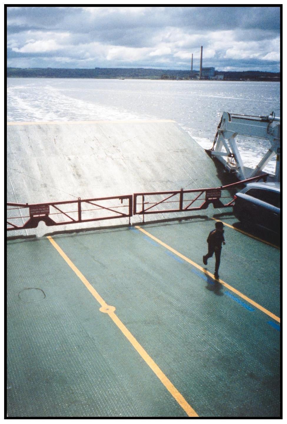 A child runs across the deck of a ferry with a body of water and industrial buildings in the background.
