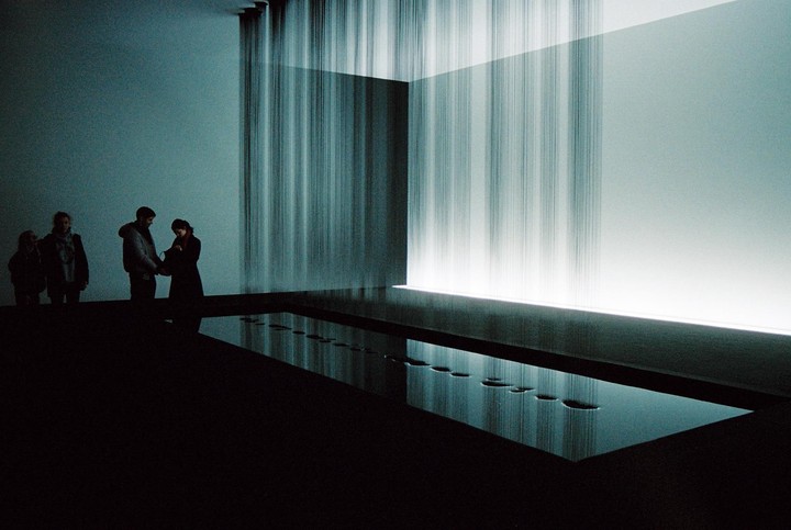 Four people stand in a dark room with a glowing wall and a pool of water reflecting light.