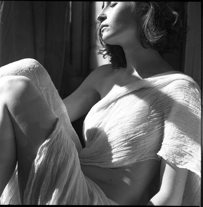 A woman with closed eyes is draped in white fabric, with sunlight casting shadows on her body.