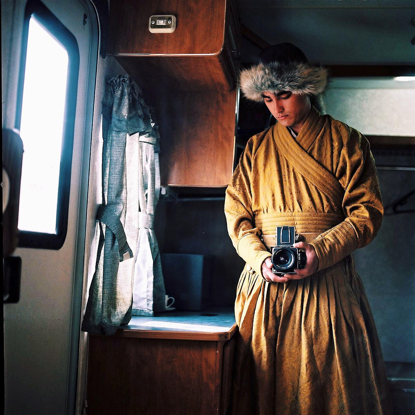 A man in a fur hat and robe holds a camera in a camper van.
