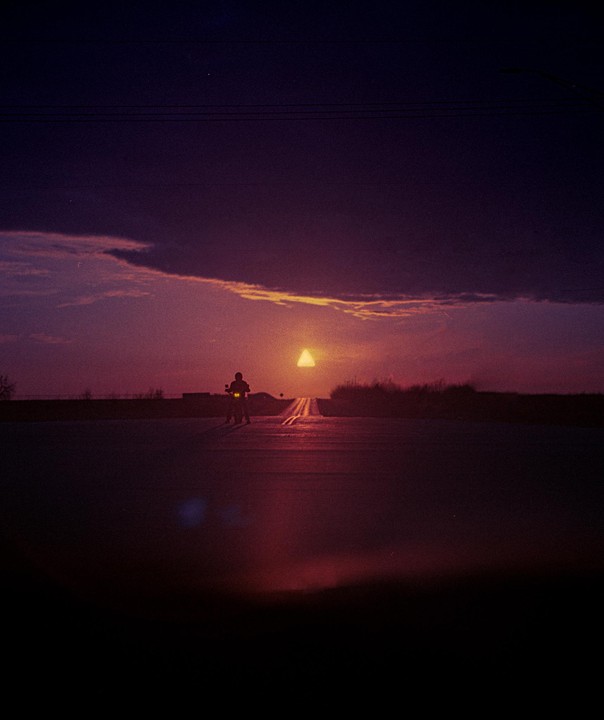 A person on a motorcycle is silhouetted against a triangular sunset on a road.