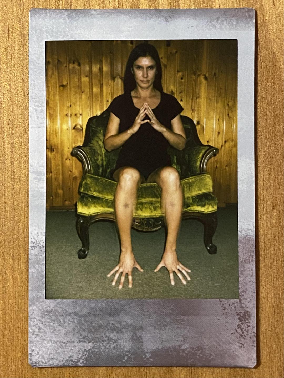 A woman sits on a green velvet armchair with her hands clasped and her feet splayed out.