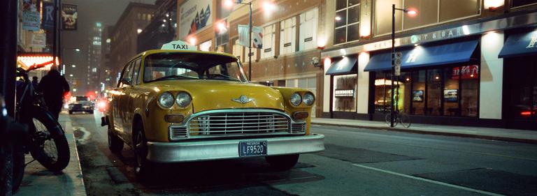 A yellow taxi cab is parked on a city street at night with buildings and streetlights in the background.