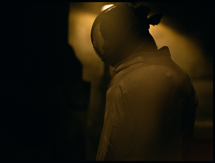 A person wearing a fencing mask and jacket stands in a dimly lit room.