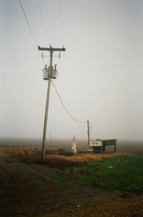 A utility pole with transformers stands in a foggy, desolate landscape next to a small structure and a dirt road.