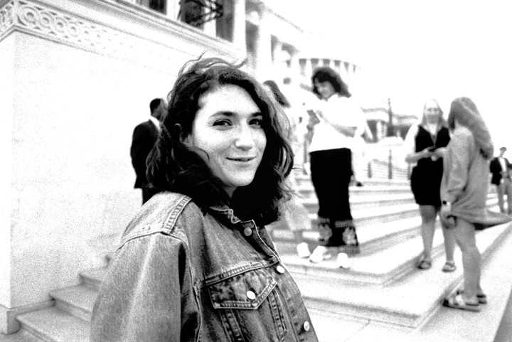 A woman in a denim jacket smiles at the camera while standing on steps with other people in the background.