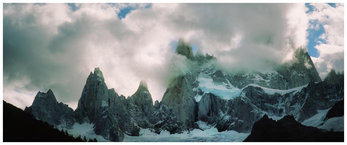 Clouds partially obscure a range of jagged, snow-capped mountains under a dramatic sky.