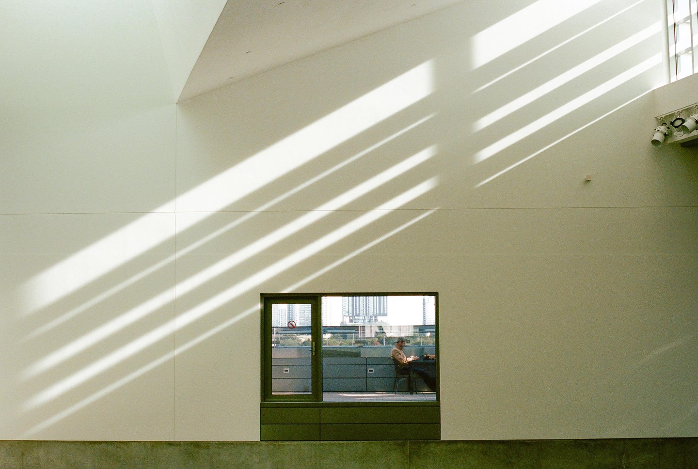 A man sits at a table outside a window, while diagonal shadows fall across the white wall inside.