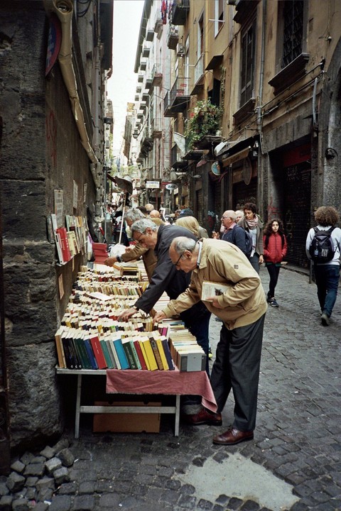 Several men are browsing books on a table in a narrow street lined with tall buildings.