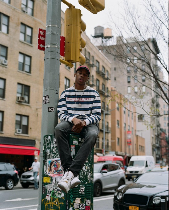 A man wearing a striped shirt and a red cap sits on a utility box on a city street.