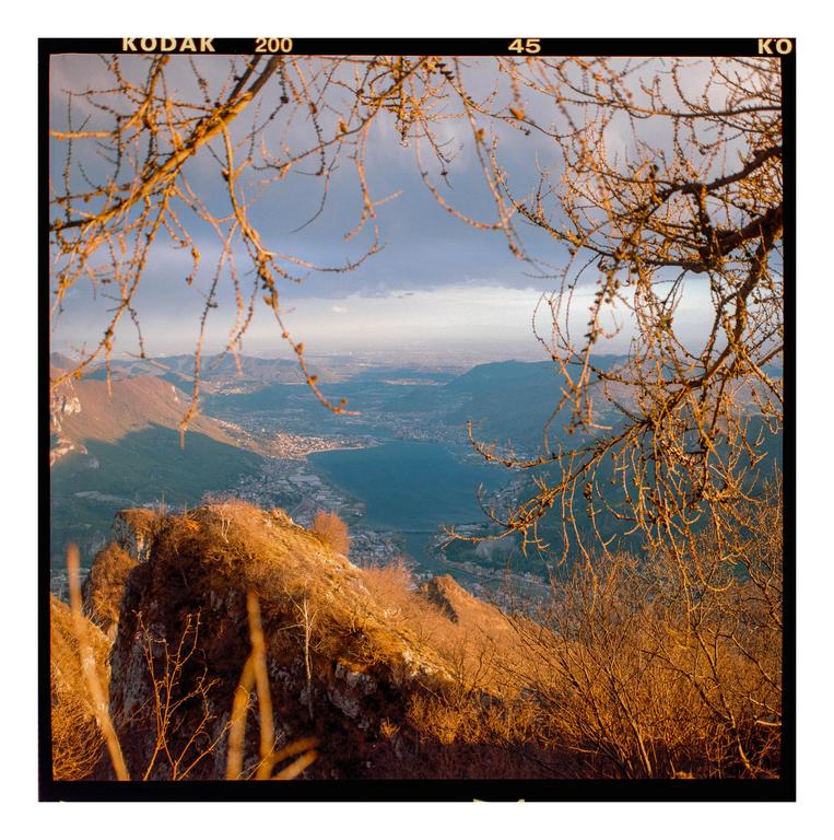 A panoramic landscape shows a lake and a town nestled between mountains, framed by bare tree branches.