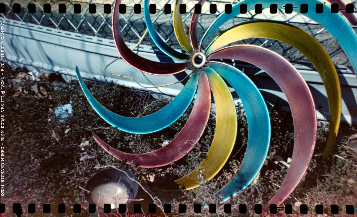 A colorful metal pinwheel stands in a garden with a chain-link fence in the background.