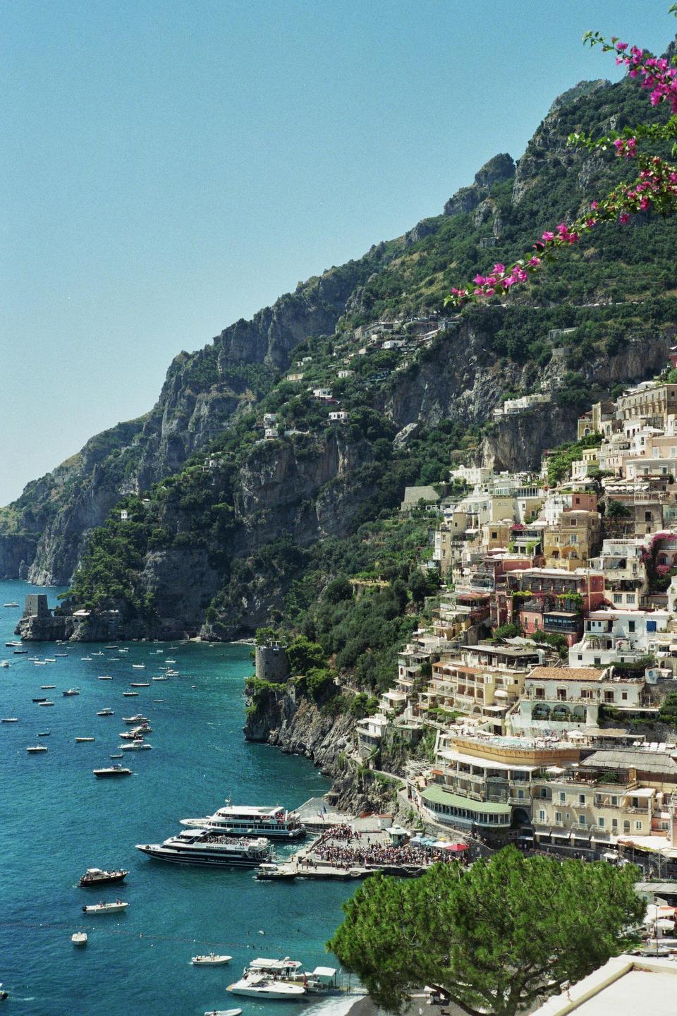 A coastal town with colorful buildings is nestled into a steep, green mountainside overlooking a bay filled with boats.