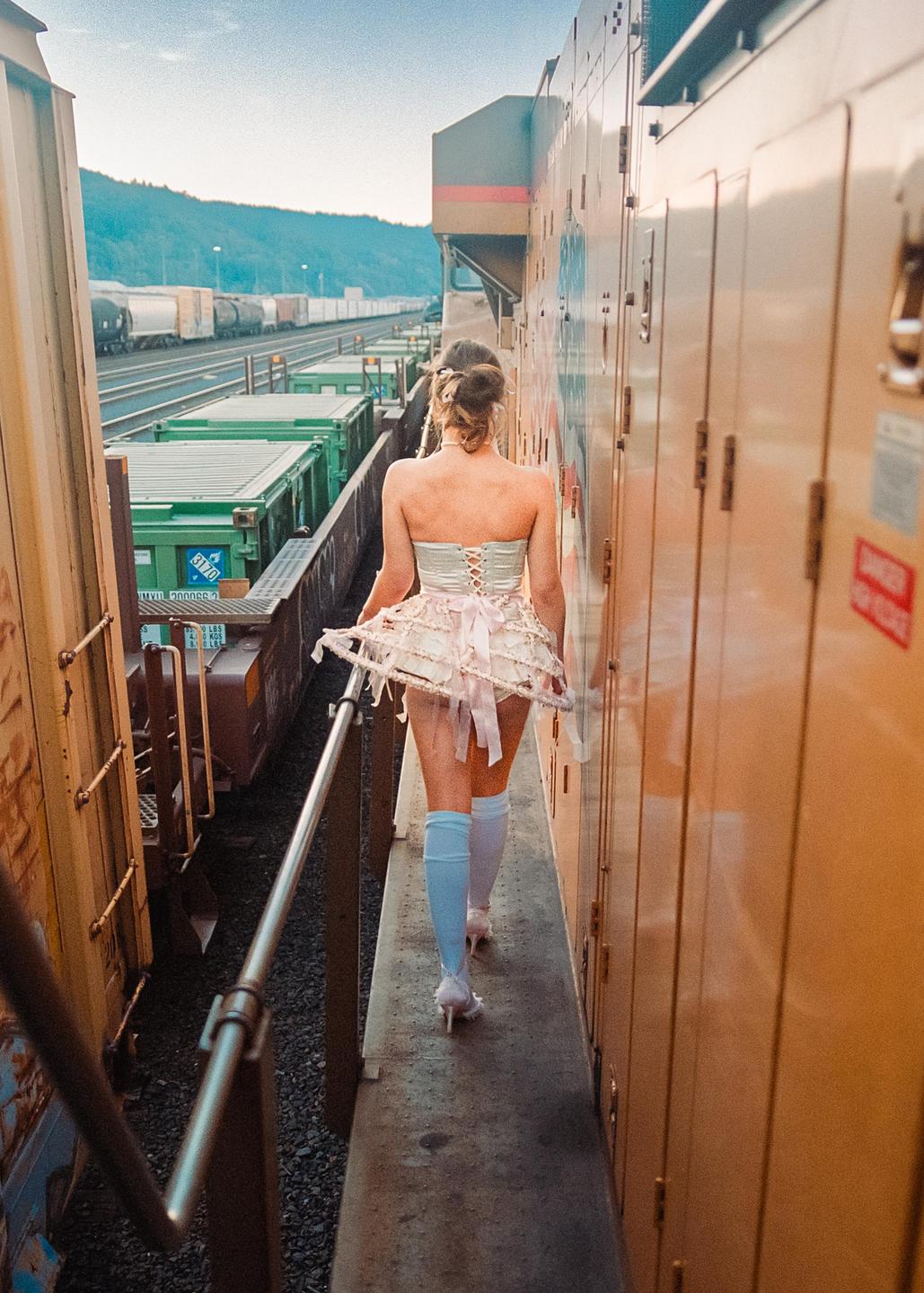 A woman in a corset and tutu walks along the side of a train.