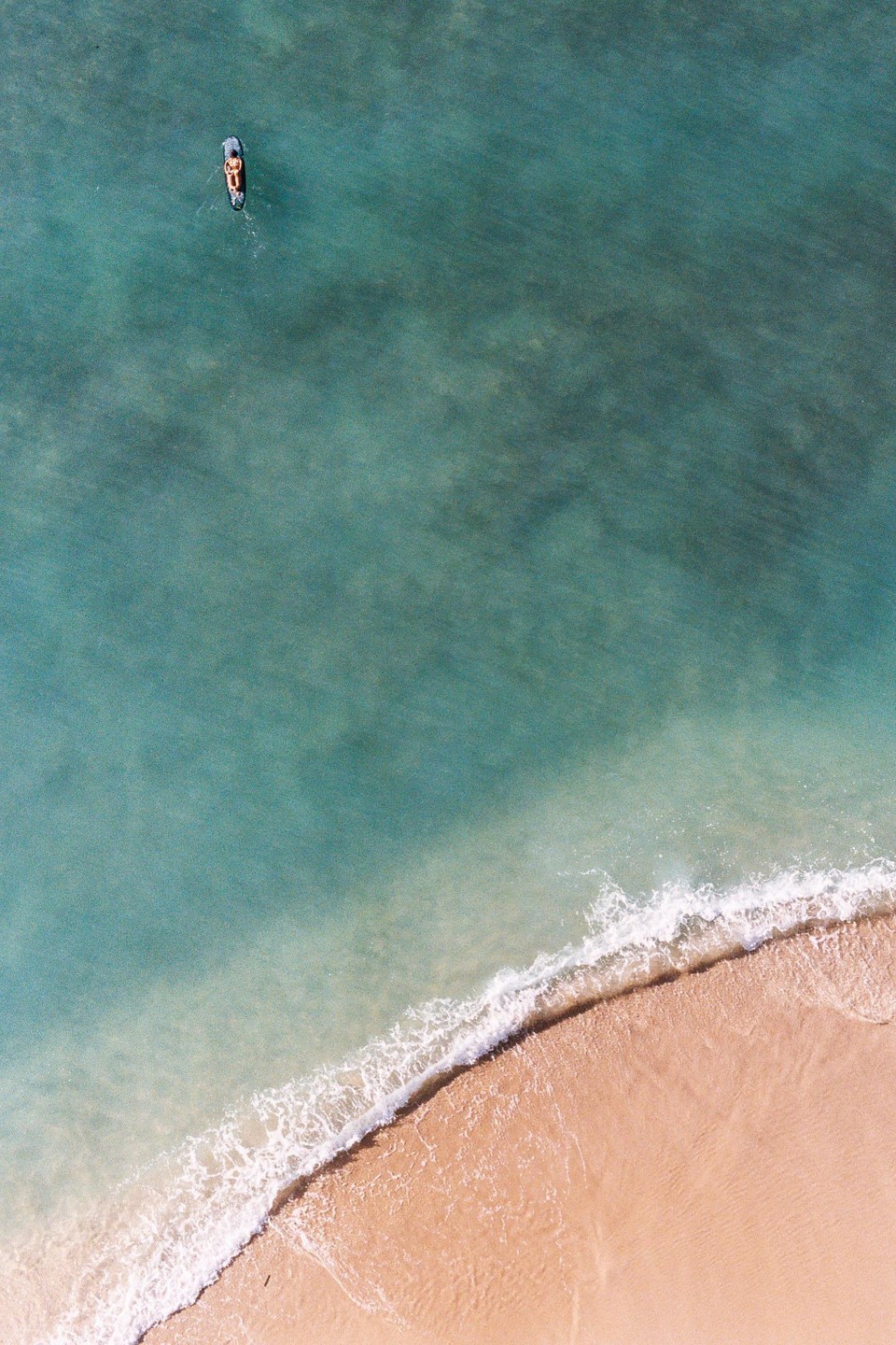 A person on a surfboard floats in turquoise water near a sandy beach with a breaking wave.