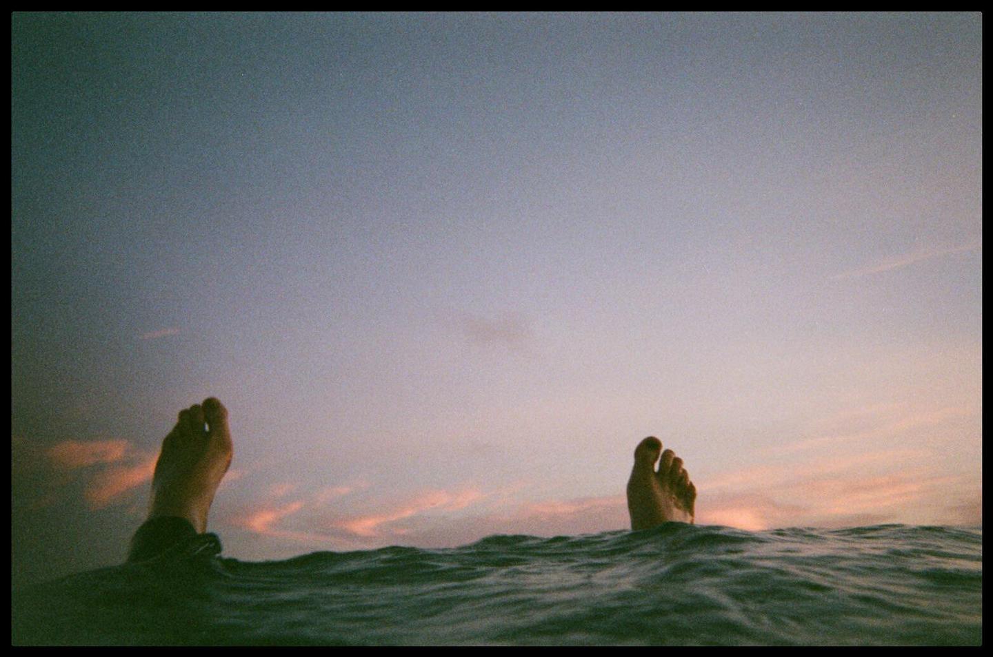 Two feet emerge from the water against a pastel sky with hints of pink clouds.