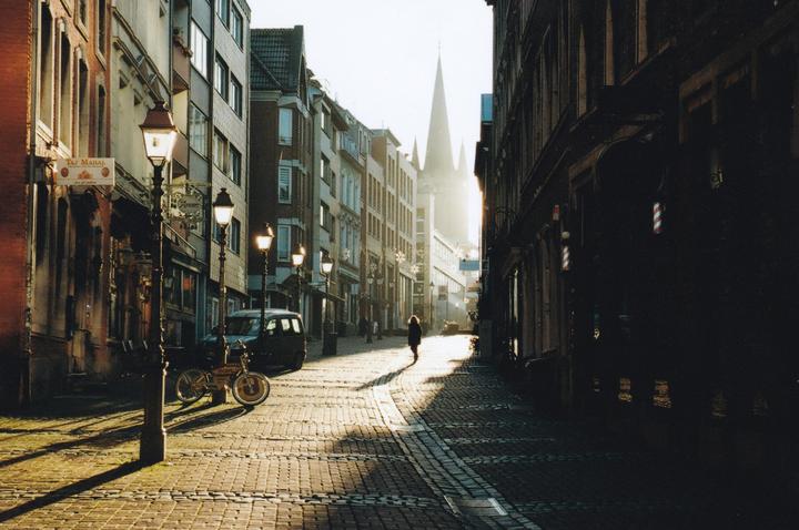 A person walks down a cobblestone street lined with buildings and streetlights, with a church steeple visible in the distance.
