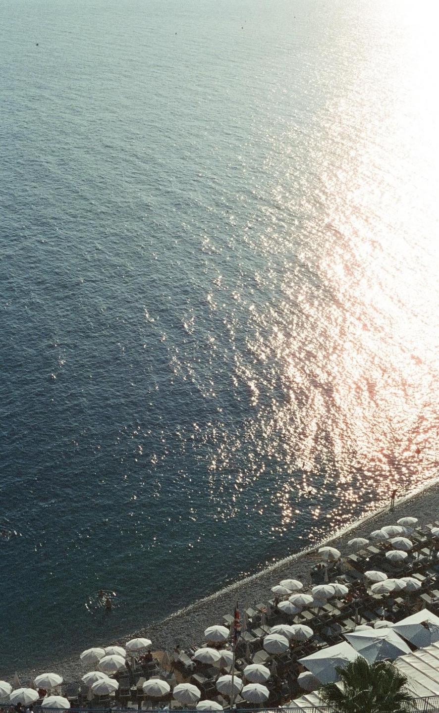 An aerial view shows a beach with many white umbrellas next to the sparkling sea.