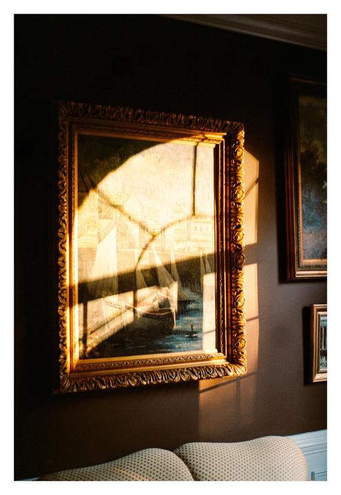 Sunlight streams through a window, casting shadows on a framed painting of boats on the water.