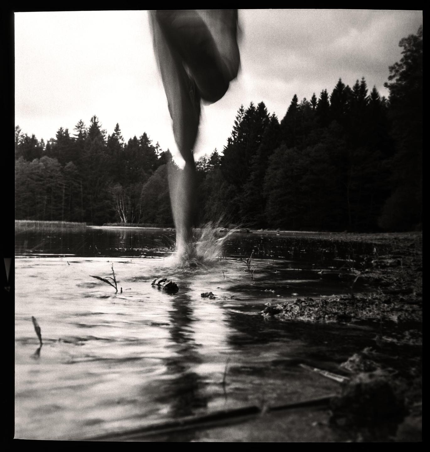 A person's legs splash into a body of water with a forest in the background.