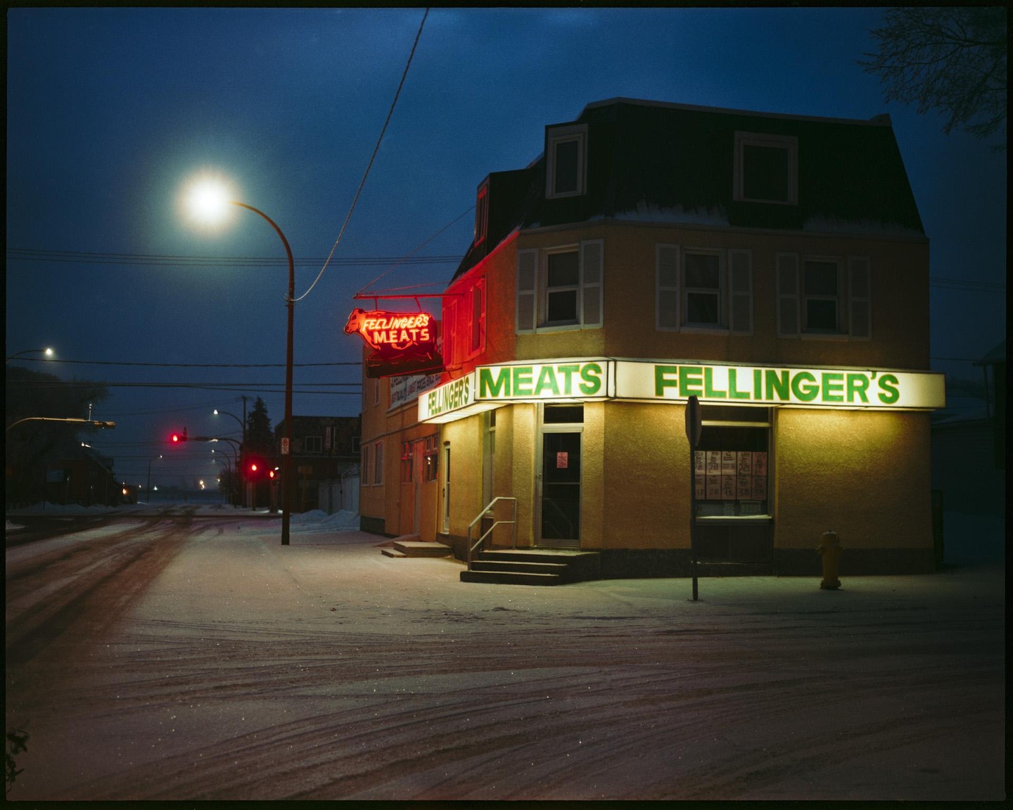 A snow-covered street corner at night features a building with illuminated signs for Fellinger's Meats.