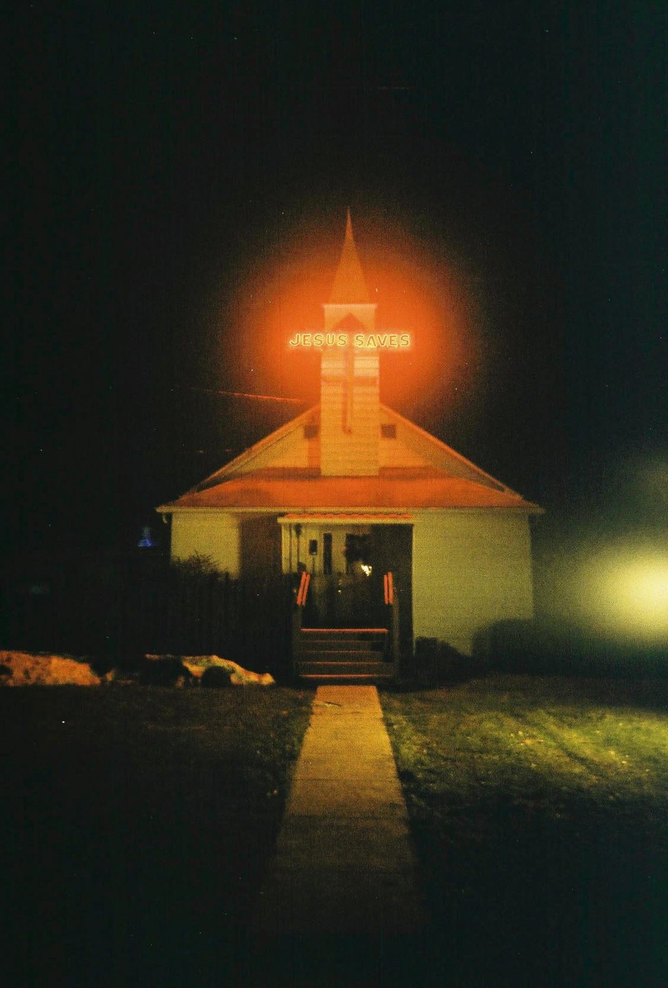 A church with a steeple and a glowing orange "Jesus Saves" sign stands at night.