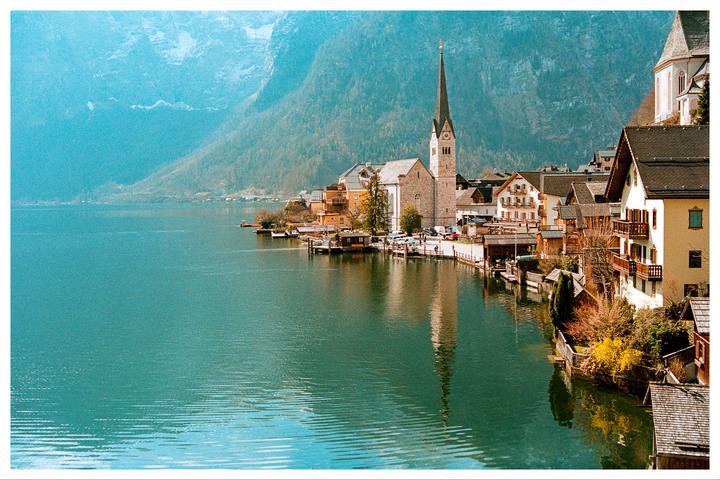 A village with a prominent church steeple is nestled beside a lake, with mountains rising in the background.