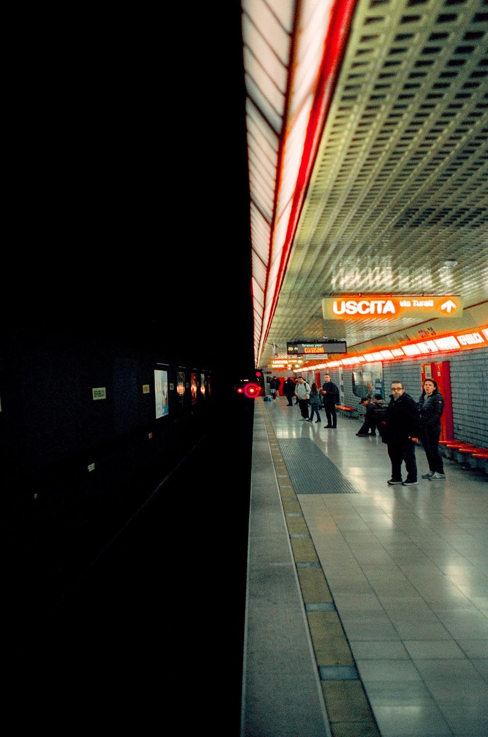 People wait on a subway platform with bright orange lights and an "USCITA" sign.