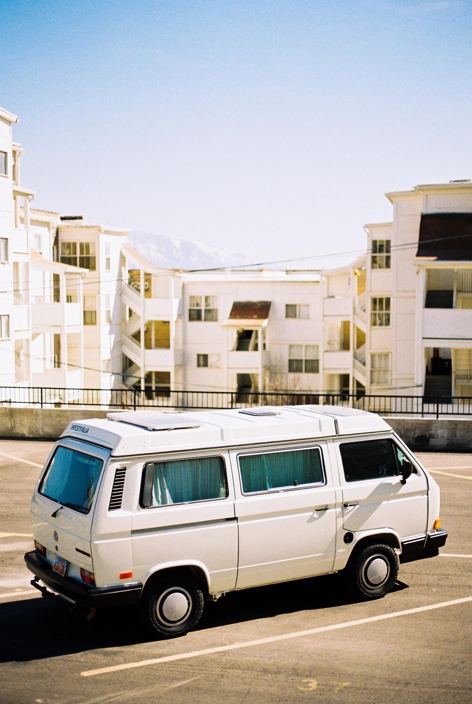 A white Westfalia van is parked in a lot with white buildings and a snowy mountain in the background.