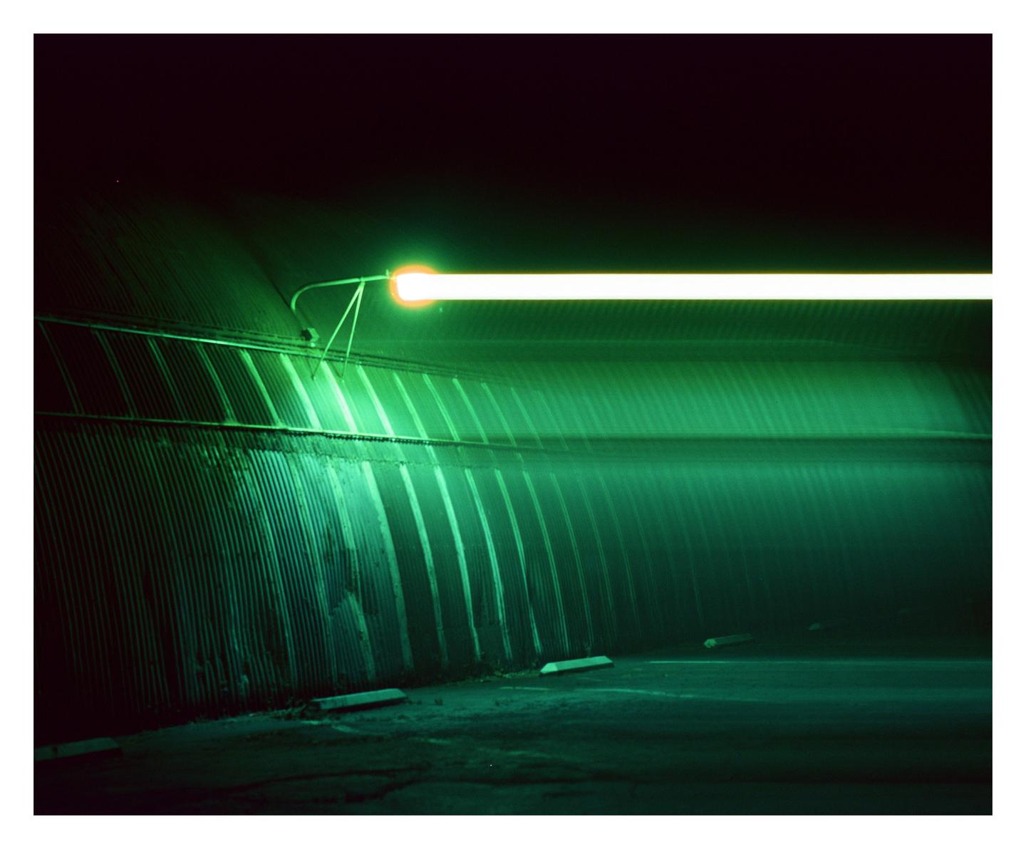 A long exposure photograph shows a bright light source illuminating a corrugated metal tunnel in green.